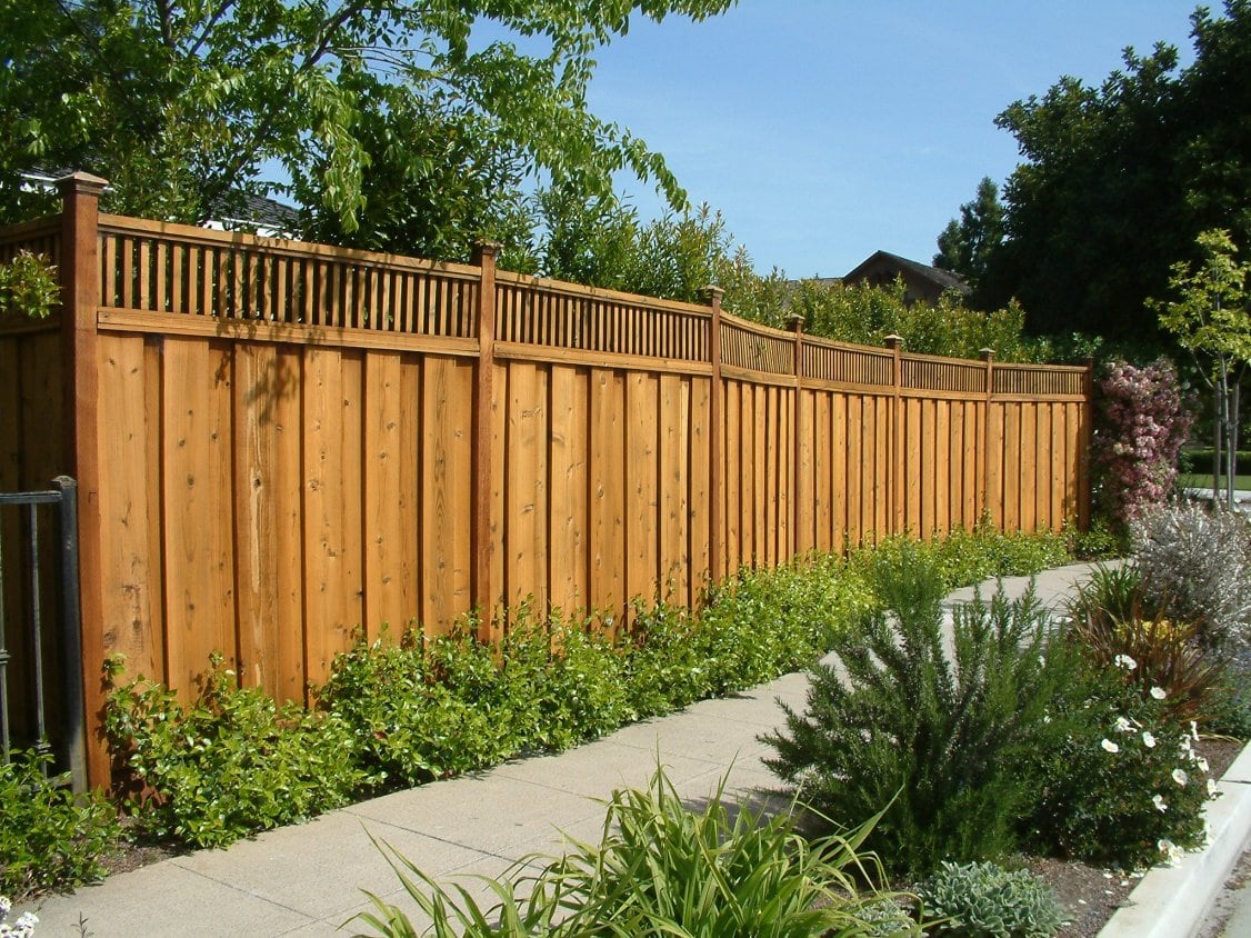 A newly installed wooden backyard fence viewed from the sidewalk. The fence is made of wooden panels that are evenly spaced and have a warm, natural tone. The fence features a decorative lattice top with square cutouts, adding a touch of elegance to the design. The fence stands tall and sturdy, providing privacy and security for the backyard. Beyond the fence, the green foliage of trees and shrubs can be seen, creating a peaceful and inviting atmosphere.