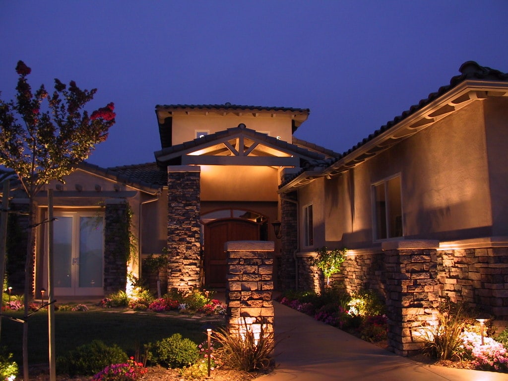 A charming front view of a house illuminated by elegant accent lighting. The warm glow of the lights highlights the unique features of the house. The landscaping is also illuminated, with light softly highlighting the greenery and flowers surrounding the house. The effect is a beautiful and cozy ambiance that makes the house feel like a welcoming home.