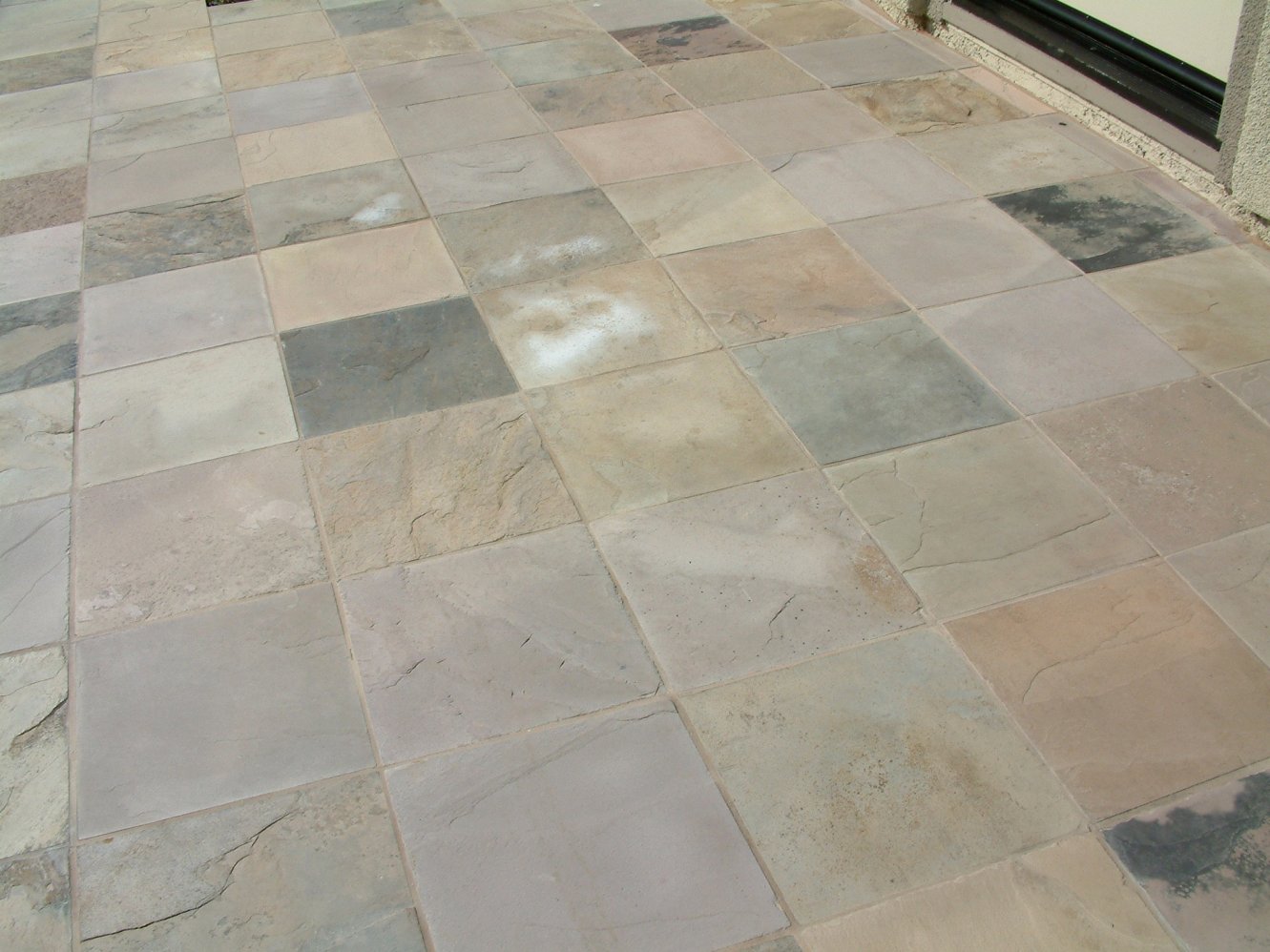 Square pavers arranged in a neat pattern on a backyard patio, surrounded by a lush green lawn and potted plants. The pavers are made of grey stone with a slightly rough texture and a matte finish, creating a rustic and natural look. The patio provides a perfect spot for outdoor entertaining, with ample seating options and a cozy fire pit in the center.