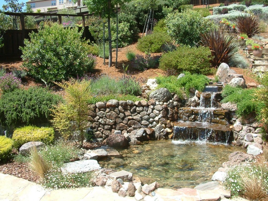 A stunning tiered backyard with a cascading stone waterfall and lush landscaping. The waterfall flows gently from the top tier to the lower tiers, creating a soothing sound and a calming atmosphere. The tiers are made of natural stone and are surrounded by vibrant greenery, including trees, shrubs, and flowers.